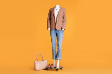Beyaz tişört, bej ceket ve turuncu arka planda kot pantolon giymiş aksesuarlı kadın mankeni. Şık kıyafet.