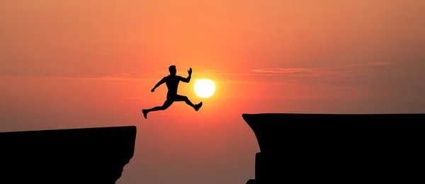 stock image Concept of reaching life and business goals. Silhouette of man jumping over chasm at sunrise