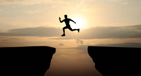 Concept of reaching life and business goals. Silhouette of man jumping over chasm at sunrise