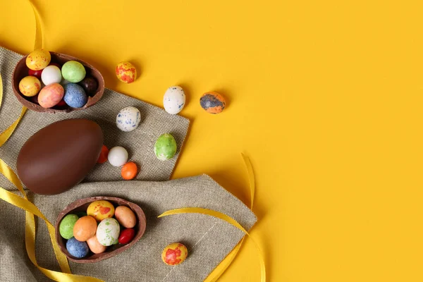 stock image Whole and halves of chocolate eggs with colorful candies on yellow background, flat lay. Space for text