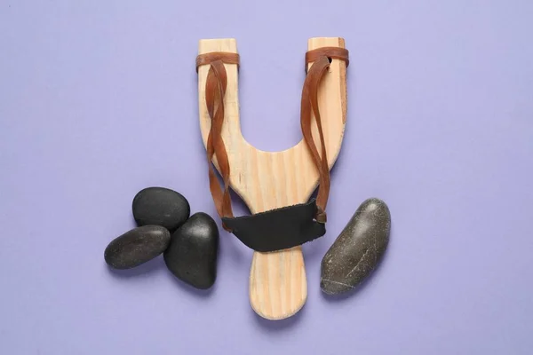 stock image Wooden slingshot with stones on violet background, flat lay