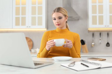 Ev işyerinde. Bir fincan sıcak içecekle mutfaktaki mermer masada dizüstü bilgisayara bakan bir kadın.
