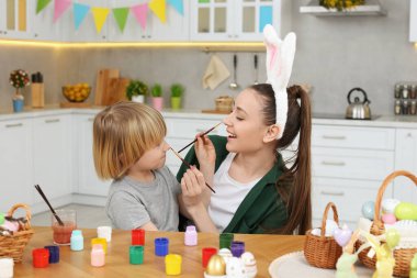 Mother and her son having fun while painting Easter eggs at table in kitchen