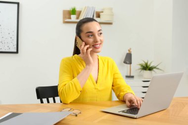 Ev işyerinde. Odanın ahşap masasında bilgisayarın yanında akıllı telefondan konuşan mutlu bir kadın.