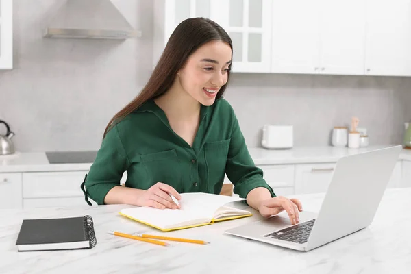 家庭工作场所在厨房的大理石办公桌前 一个快乐的女人带着笔和笔记本在笔记本电脑上工作 — 图库照片
