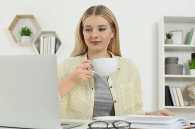 Home workplace. Woman with cup of hot drink working on laptop at white desk in room