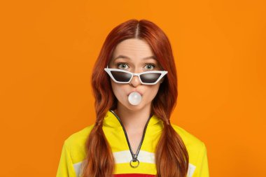 Güneş gözlüklü bir kadının portresi turuncu arka planda sakız üflüyor.