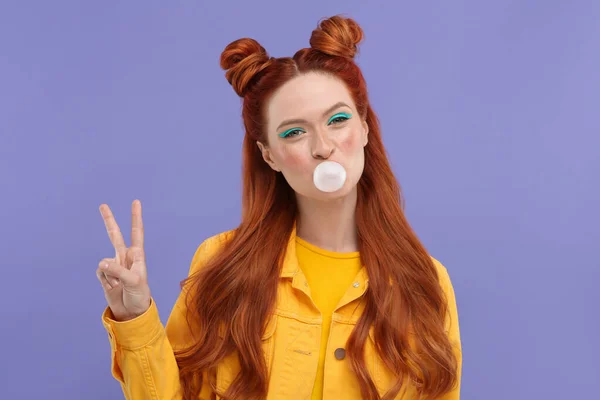 stock image Portrait of beautiful woman with bright makeup blowing bubble gum and showing peace gesture on violet background