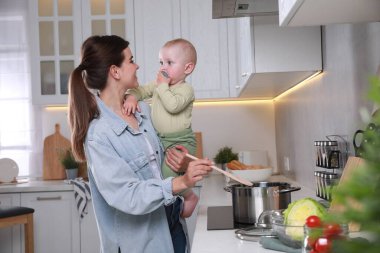 Mutfakta yemek pişirirken küçük bebeğini kucağında tutan mutlu genç kadın.