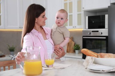 Mutfakta meyve suyu içerken küçük bebeğini kucağında tutan mutlu genç bir kadın. Mesaj atmak için yer var.