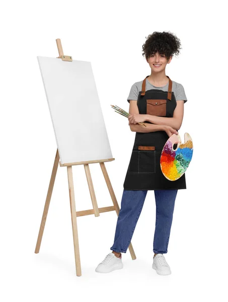 年轻女子在画架附近拿着画笔和艺术家的调色板 画架上有白色背景的帆布 — 图库照片