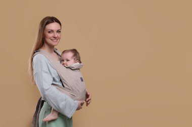 Anne, açık kahverengi arka planda çocuğunu askıda (bebek taşıyıcısı) tutuyor. Metin için boşluk