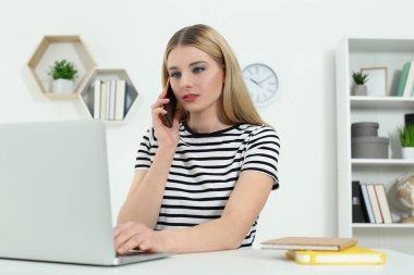 Ev işyerinde. Beyaz masadaki bilgisayarın yanında akıllı telefondan konuşan bir kadın.