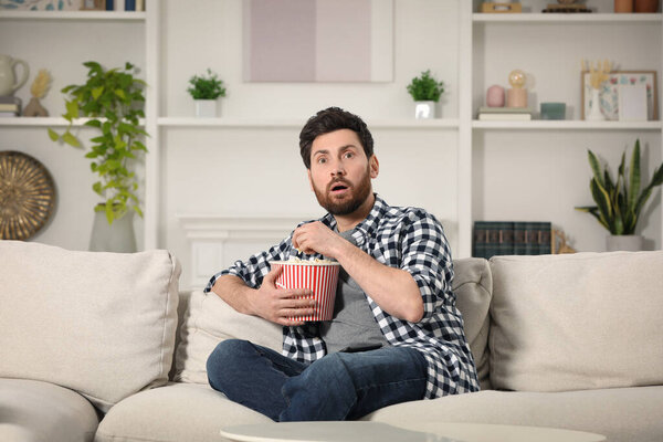 Surprised man watching TV with popcorn on sofa at home