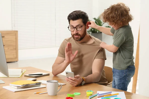 stock image Little boy bothering his father at home. Man working remotely at desk