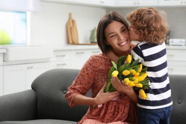 Little son kissing and congratulating his mom with Mother`s day at home, space for text. Woman holding bouquet of yellow tulips