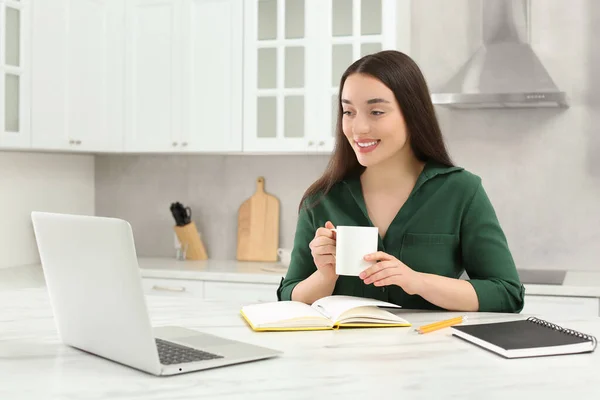 家庭工作场所快乐的女人 一边喝着热饮 一边看着厨房里大理石桌子旁的笔记本电脑 — 图库照片