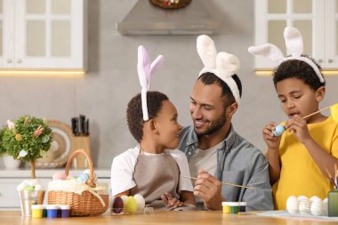 Mutlu Afrikalı Amerikalı baba ve tatlı çocukları mutfaktaki masada Paskalya yumurtası boyuyor.