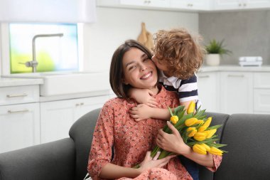 Little son kissing and congratulating his mom with Mother`s day at home. Woman holding bouquet of yellow tulips
