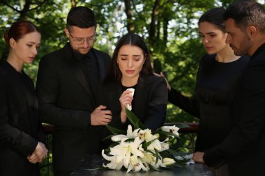 Cenaze töreni. Granit mezar taşının yanında yas tutan üzgün insanlar ve mezarlıkta beyaz zambaklar.