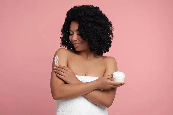 Young woman applying body cream onto shoulder on pink background