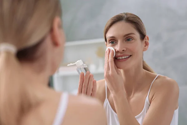 stock image Beautiful woman removing makeup with cotton pad near mirror indoors