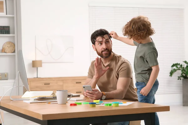 stock image Little boy bothering his father at home. Man working remotely at desk