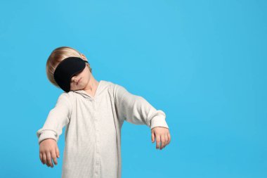 Boy in pajamas and sleep mask sleepwalking on light blue background, space for text