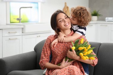 Little son kissing and congratulating his mom with Mother`s day at home. Woman holding bouquet of yellow tulips