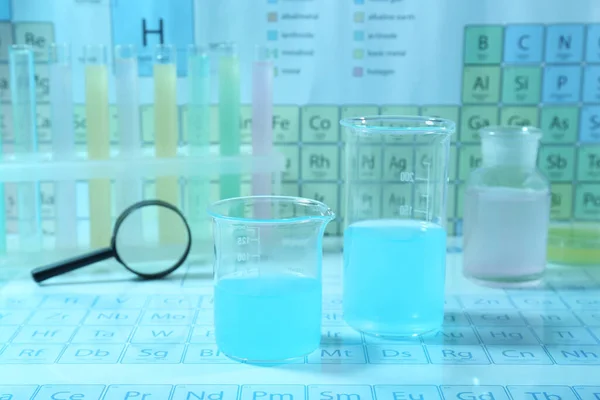 不同的实验室玻璃器皿 装有液体和放大镜 放在化学元素的周期表上 浅蓝色调效果 — 图库照片