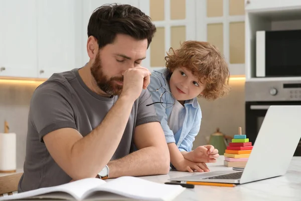 彼は家でリモートで働いている間 父親を悩ませている小さな男の子 ノートパソコンを持つ男と台所の机の上に彼の子供 — ストック写真