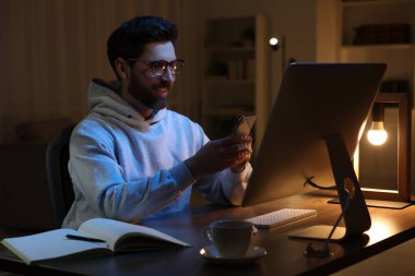 Ev işyerinde. Geceleri odasındaki ahşap masada bilgisayarla çalışırken akıllı telefon kullanan mutlu bir adam.