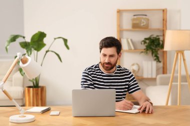 Ev işyerinde. Odanın ahşap masasında dizüstü bilgisayarla çalışırken not alan bir adam.