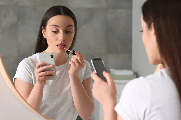 屋内で鏡の近くに口紅を適用しながら スマートフォンを使用して美しい若い女性 インターネット中毒 — ストック写真