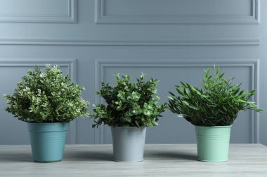 Gri duvarın yanındaki beyaz ahşap masadaki saksılarda birçok farklı yapay bitki var.