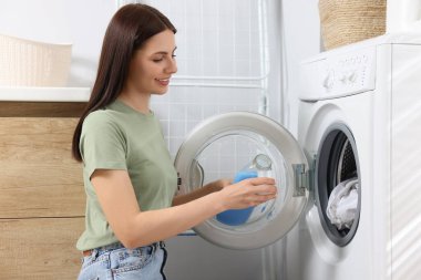 Kadın banyoda çamaşır makinesinin yanında şişeden kapağa yumuşatıcı döküyor.