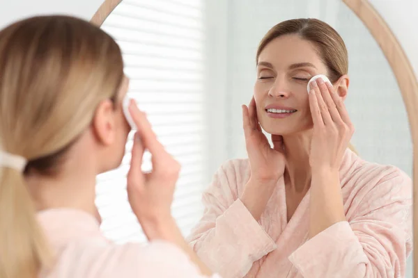 stock image Beautiful woman removing makeup with cotton pad near mirror indoors