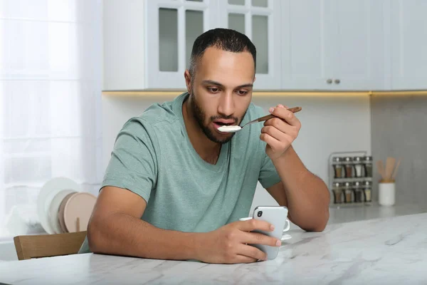 年轻人在厨房的白色大理石桌子边吃早餐时 用智能手机 上网成瘾 — 图库照片