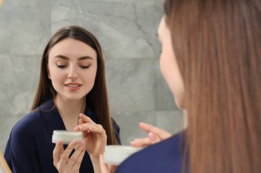 Mavi bornozlu güzel kadın aynaya bakıyor ve içeride krem sürüyor.