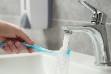 Banyodaki musluktan akan suyun altında plastik diş fırçası yıkayan kadın, yakın plan.