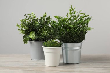 Tahta masadaki saksılarda, açık gri arka planda birçok farklı yapay bitki var.