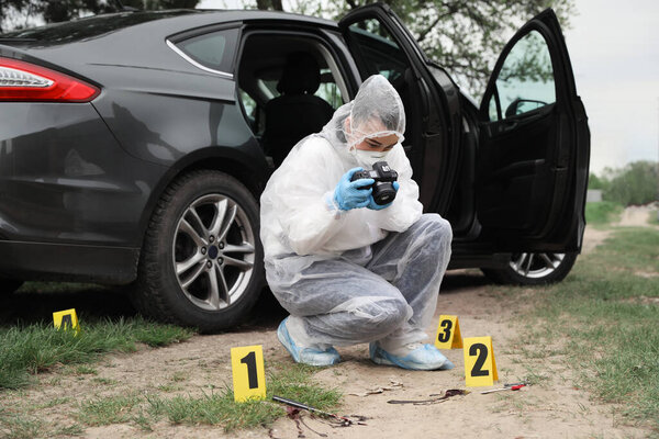 Criminologist in protective suit taking photo of evidences at crime scene outdoors