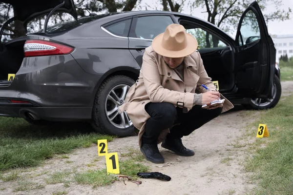 Professional detective in hat examining crime scene near car outdoors