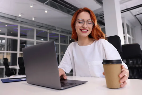 在办公室的白色桌子上 一个快乐的女人拿着纸杯咖啡在笔记本电脑上工作 — 图库照片