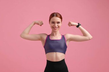 Spor kıyafetli genç bir kadın pembe arka planda kaslarını gösteriyor.
