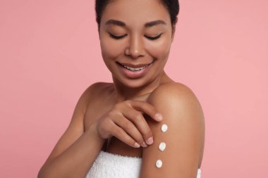 Genç bir kadın pembe arka planda vücut kremi sürüyor.