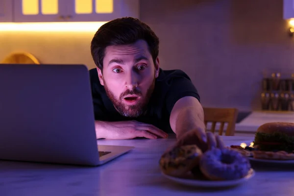 stock image Man taking donut while using laptop in kitchen at night. Bad habit