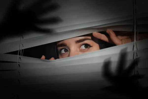 stock image Worried woman looking through window blinds into darkness. Shadow of hands with long claws. Paranoia concept