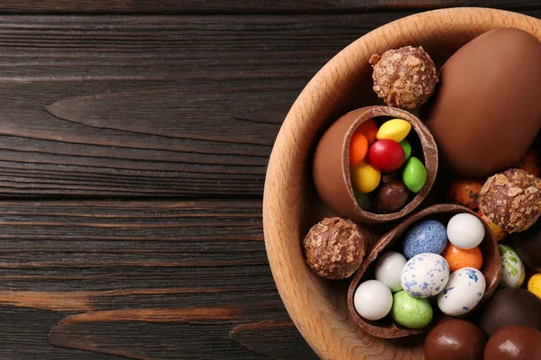 stock image Tasty chocolate eggs and sweets in bowl on wooden table, top view. Space for text