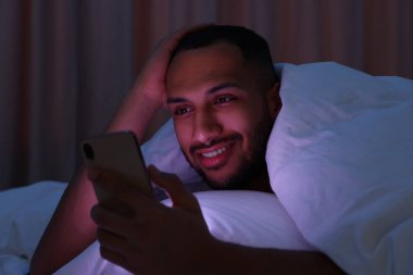 Geceleri yatakta battaniyenin altında akıllı telefon kullanan mutlu bir adam. İnternet bağımlılığı